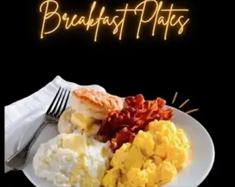 A breakfast plate with scrambled eggs, crispy bacon, grits, and a biscuit on a white plate with a fork and napkin.