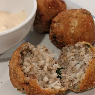 Three breaded and fried rice balls on a plate, with one cut open to show the filling. A small bowl of dipping sauce is beside them.