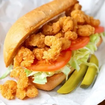 A sandwich filled with fried shrimp, lettuce, and tomato on a bun, served with pickle slices on the side.