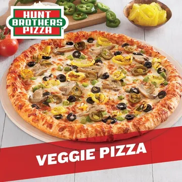 A veggie pizza topped with mushrooms, olives, onions, and peppers. A whole tomato and sliced green peppers are in the background. Hunt Brothers Pizza logo is at the top left.